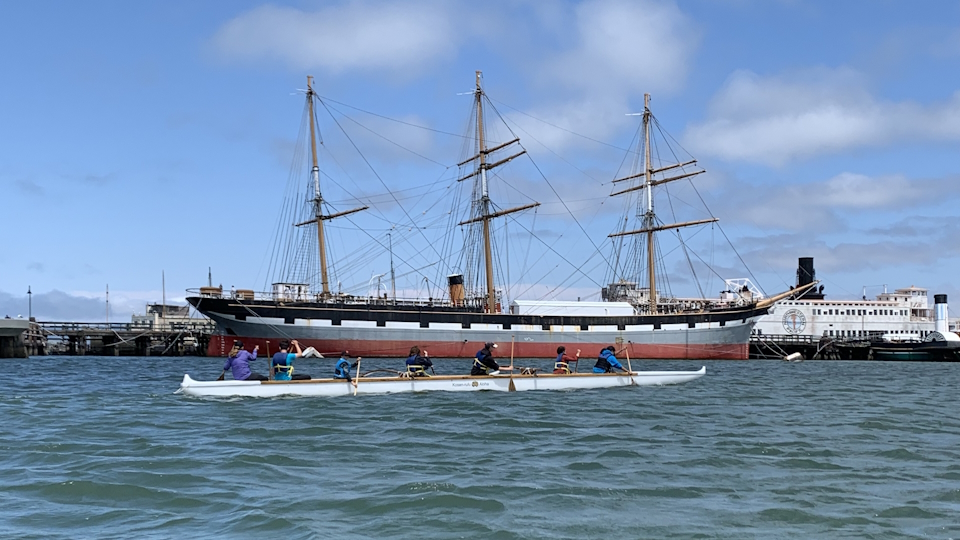 Scouts at the Maritime Explorer Club day camp paddling a long canoe with a three mast sailing ship in the background